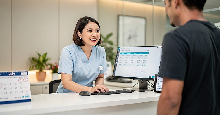 Friendly clinic receptionist smiling at reception desk with appointment scheduling workflow visible