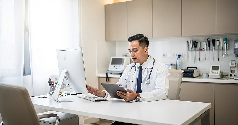 Modern Malaysian doctor reviewing patient electronic health records on a tablet in a professional clinic office with medical equipment and calm professional atmosphere.